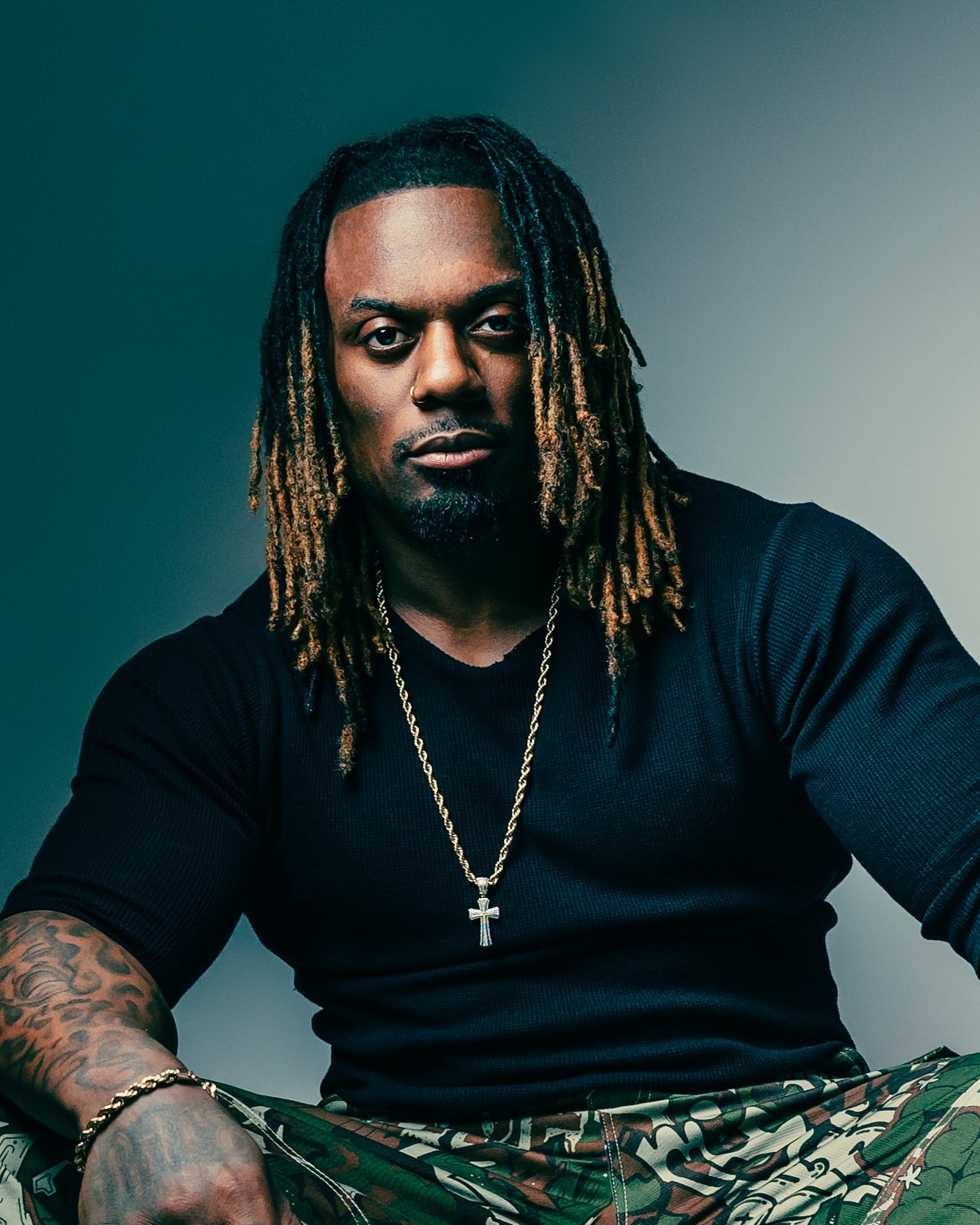 Dat Boi Jonni close portrait, direct gaze, gold cross chain, dreadlocks, moody dark studio lighting