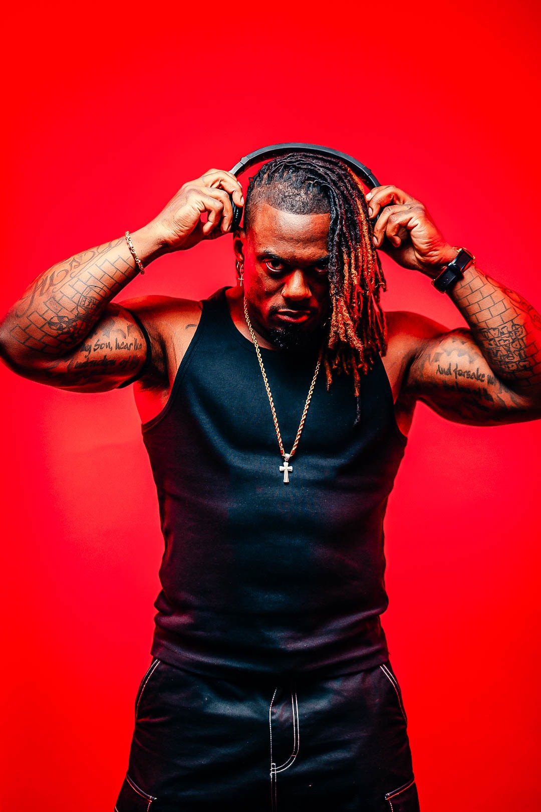 Dat Boi Jonni portrait on red background, headphones raised, tattoos visible, direct gaze
